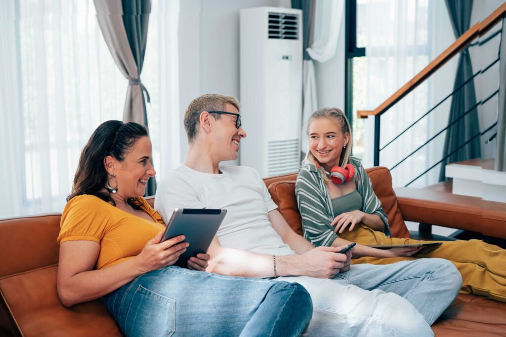 Family of three—mother, father, and teenage daughter—sitting on a living-room couch, smiling and holding tablets, with a ductless mini-split air handler visible behind them.