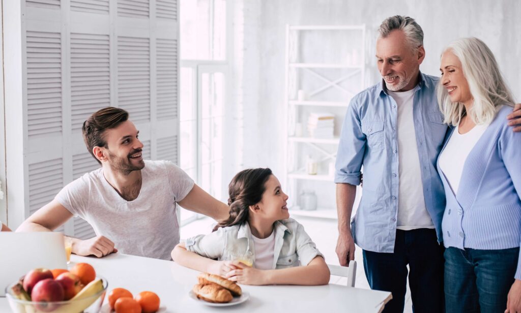 A young father and daughter seated at a kitchen table smile up at their grandparents who stand beside them, with a functional home interior in the background.